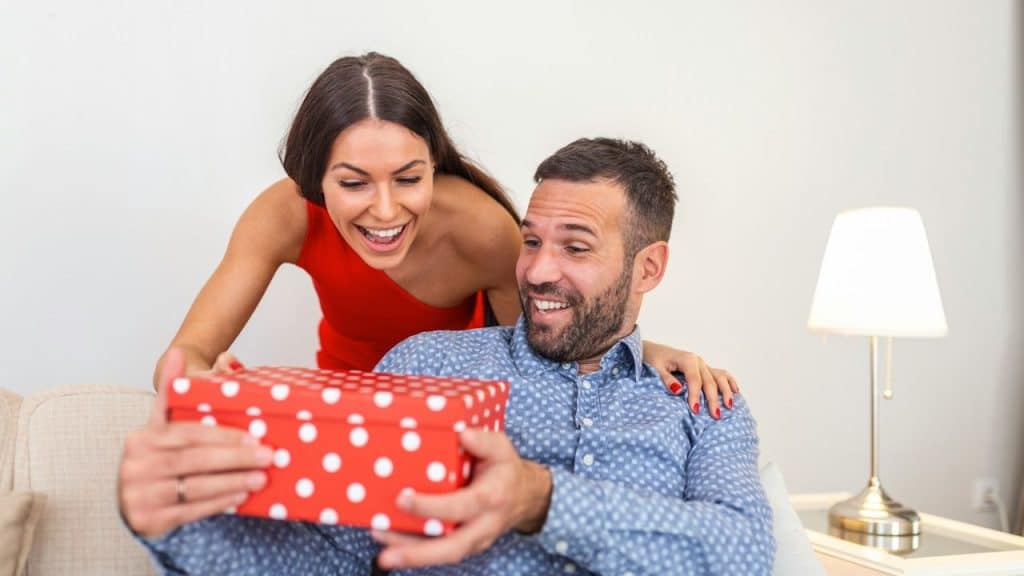 A man is smiling while opening a red polka-dot gift box, as a woman in a red dress smiles over his shoulder.