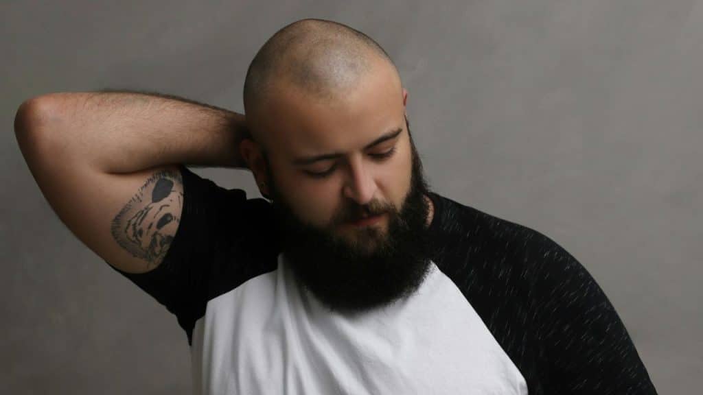 A bald, bearded man in a baseball tee stands against a gray background with his arm behind his head, looking down.