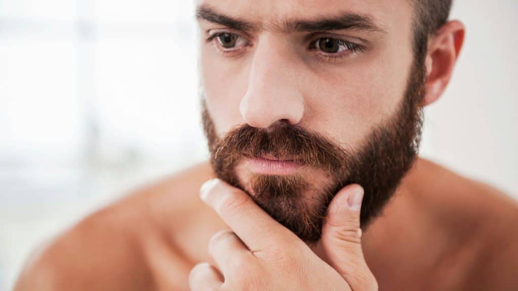A bearded man with a serious expression is touching his chin with his hand.