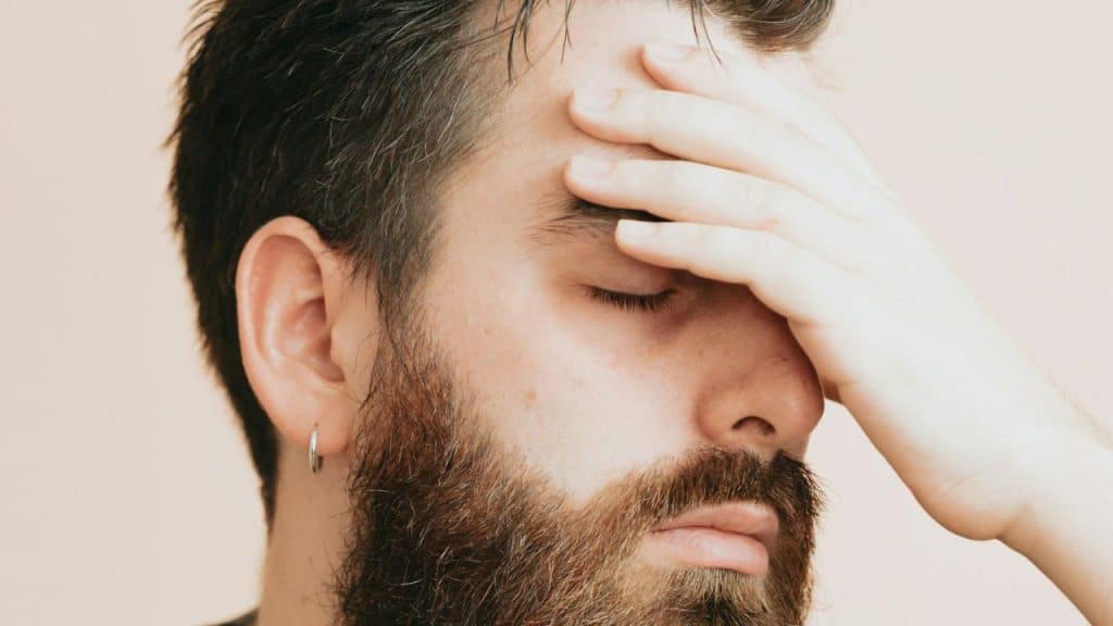 A bearded man with a silver hoop earring has his eyes closed and his hand covering his forehead.