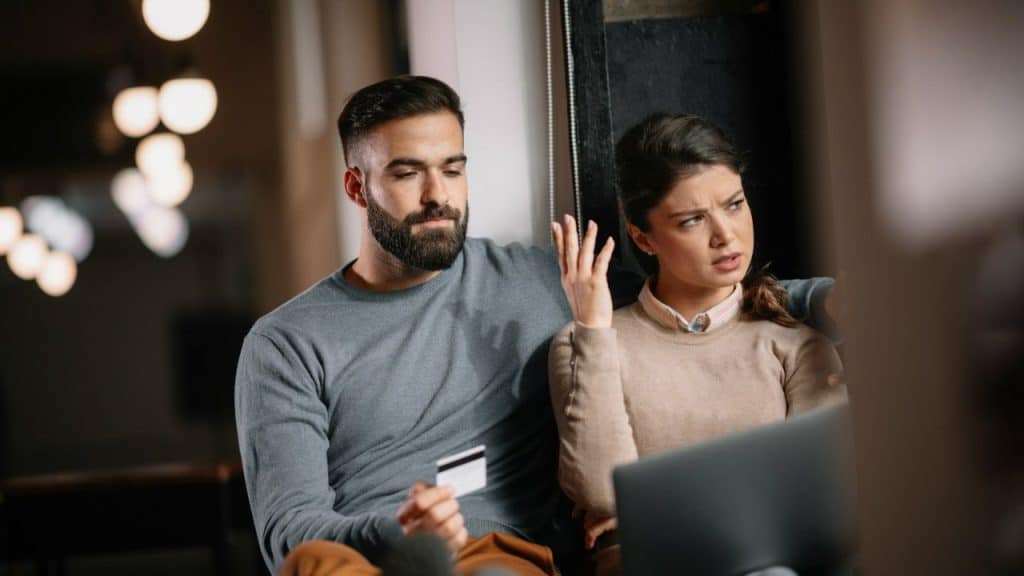 A man holds a credit card while looking at a displeased woman next to a laptop.