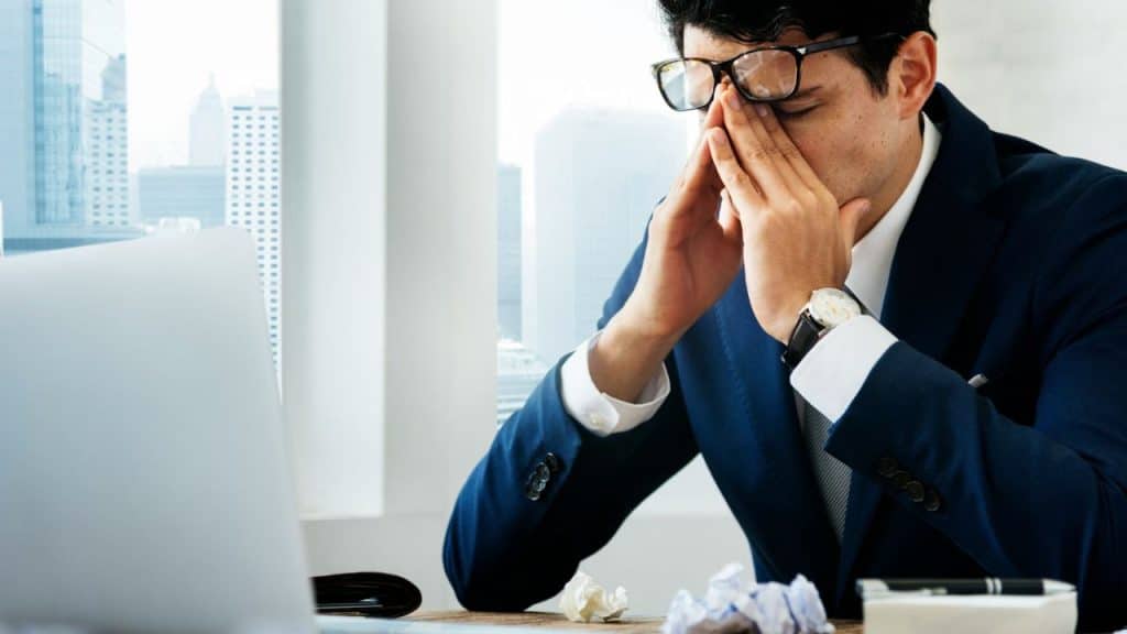 A man in a suit and glasses sits at a desk with a laptop, pressing his fingers to his eyes.