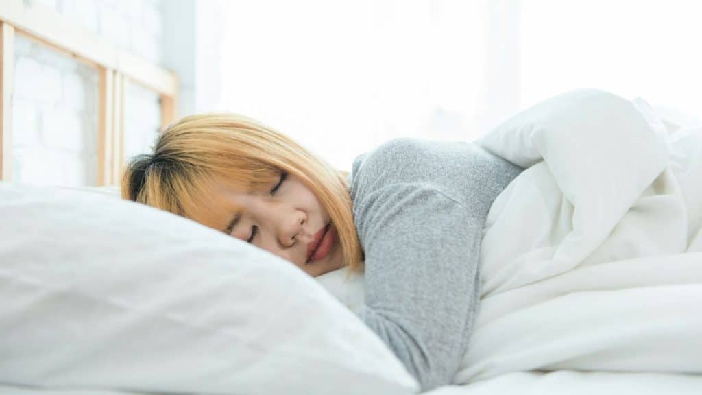 A woman with light orange hair is sleeping on her side on a white pillow in bed.