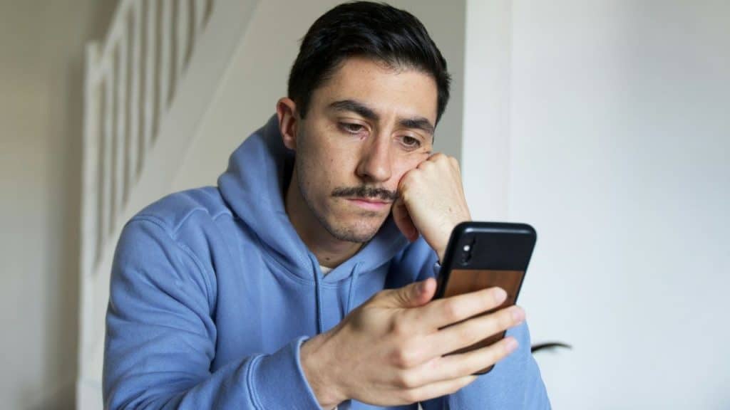 A man in a blue hoodie rests his head on his hand while looking down at his phone with a sad expression.