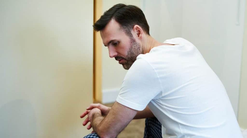 A man in a white t-shirt sits with his hands clasped, looking down sadly.
