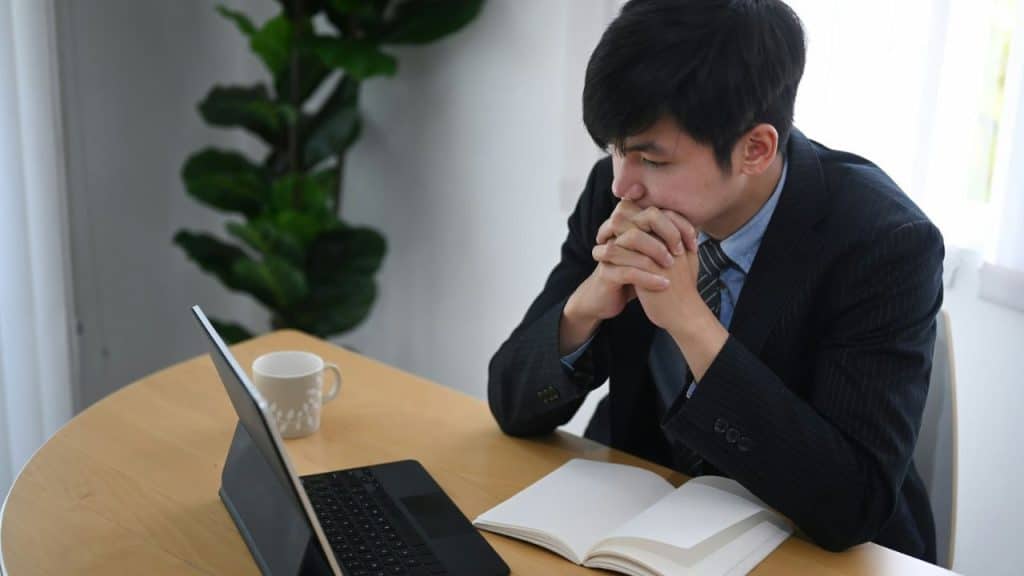 Young man in a suit sitting at a table, looking thoughtfully at a laptop and notebook.