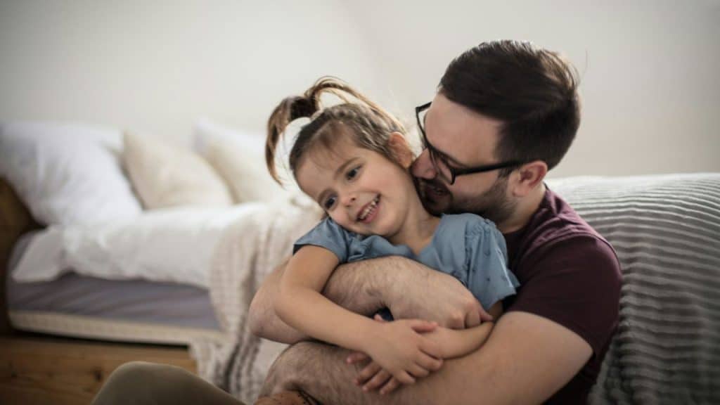 Smiling father wearing glasses hugging his young daughter in a bright, casual room.