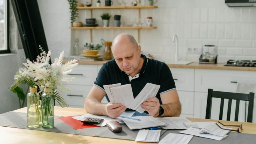 Bald man sitting at a kitchen table, intently reviewing several bills and papers.