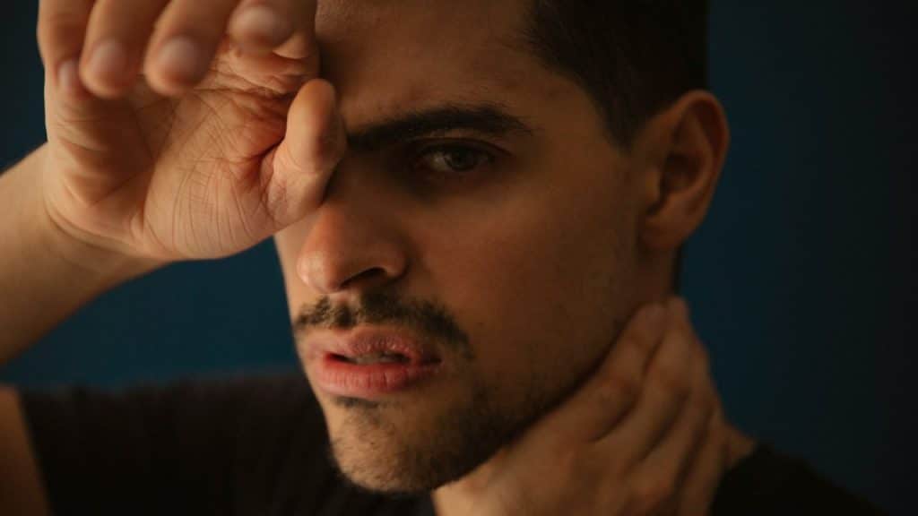 Close-up of a man with a mustache, holding his hand over his forehead and neck.