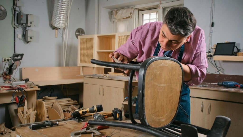 Man in a pink shirt and apron sanding the back of a wooden chair in a cluttered workshop.