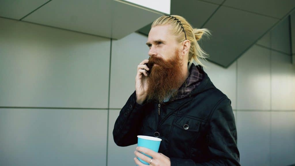 The man with long hair and unkempt beard is walking on the street while talking to someone on his phone.