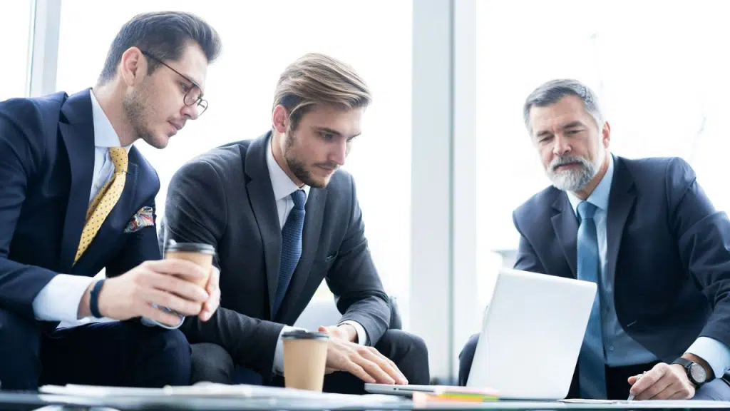 Three men in suits look intently at a laptop during a business meeting.