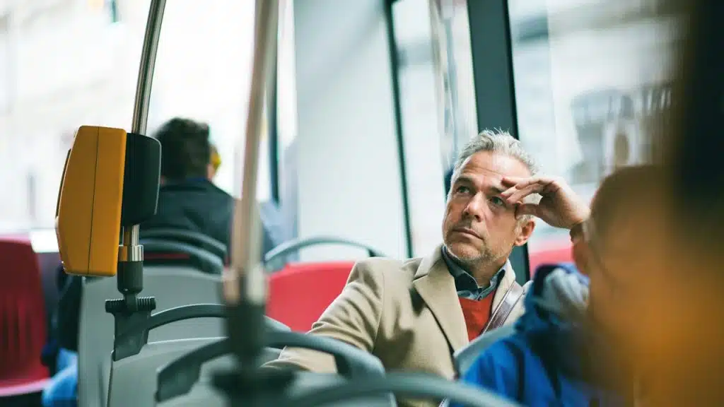 A pensive middle-aged man in a tan coat and red sweater riding a bus.