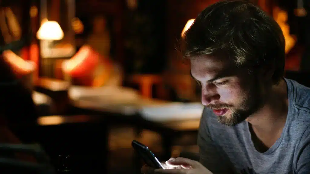 A close-up of a man in a dark, dimly lit room, illuminated by the bright screen of the smartphone he is looking at.