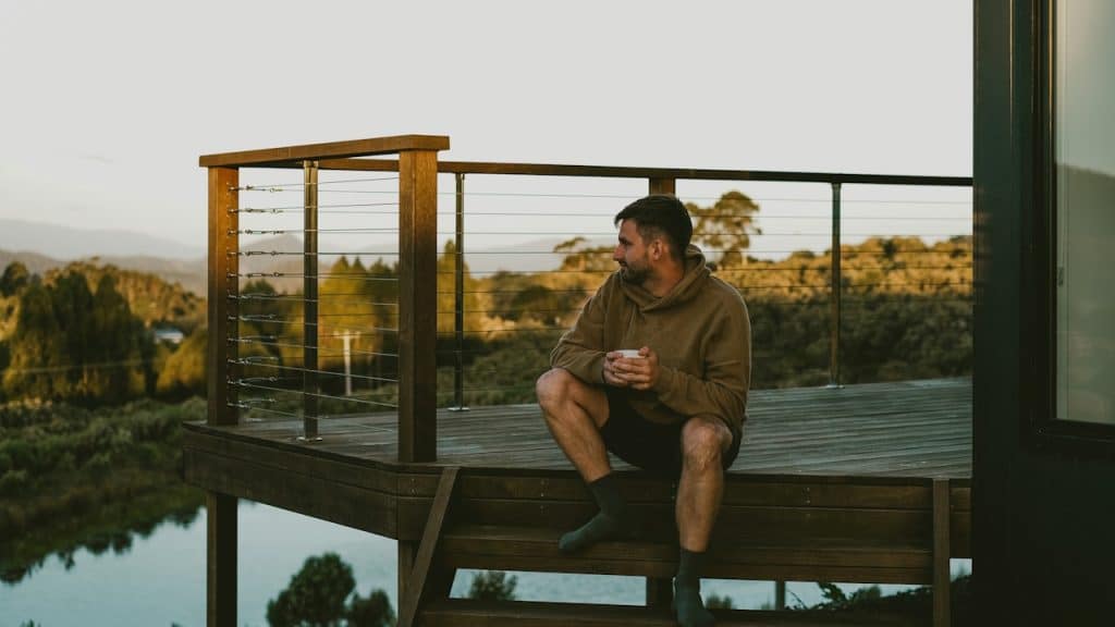 A man in a brown hoodie sits on the wooden steps of a deck, holding a mug.