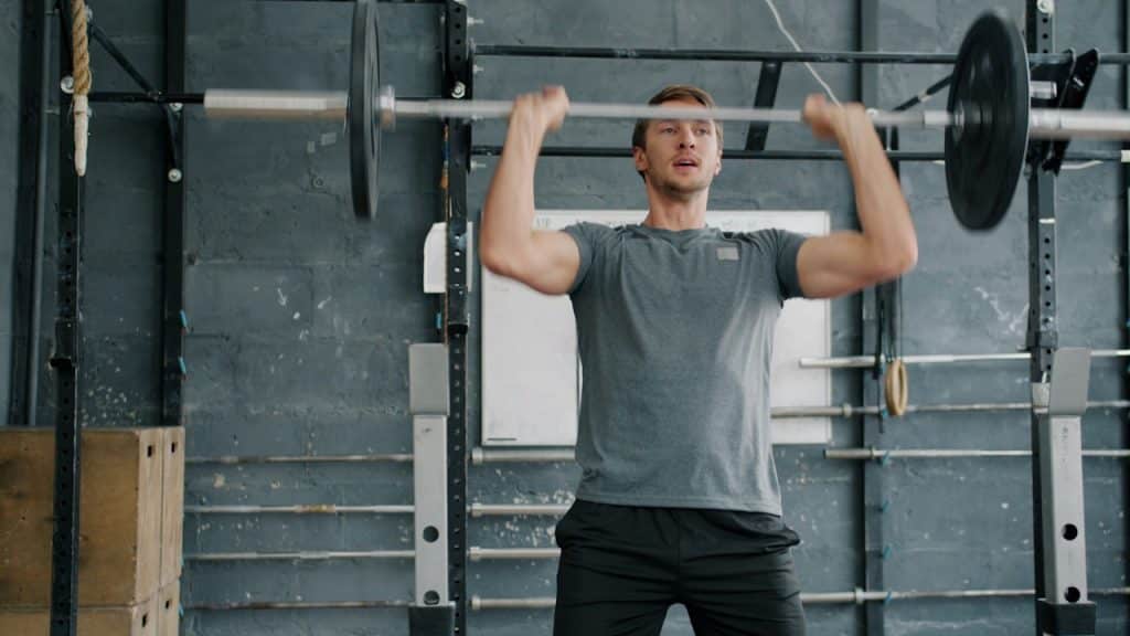 A strong, focused man in a gray t-shirt and black shorts performing an overhead press.