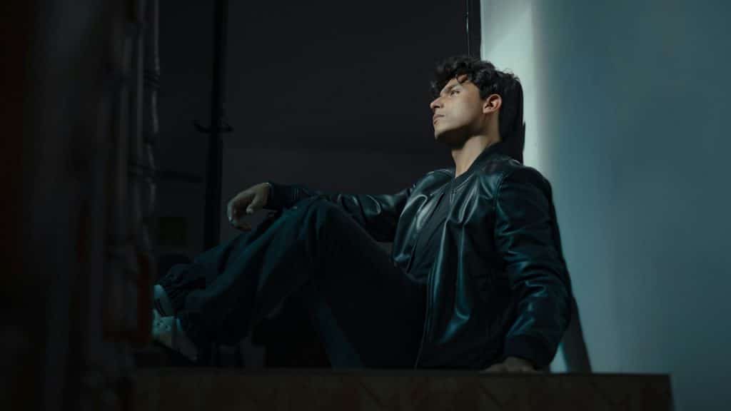 A stylish young man in a black leather jacket and dark jeans sitting on the corner of a staircase landing in the dark.