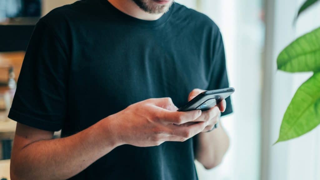A close-up of a man in a plain black t-shirt holding a smartphone with both hands while looking down.
