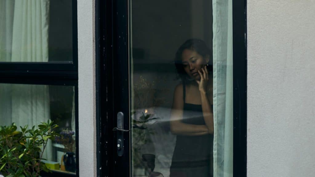 A somber-looking woman in a dark tank top is seen through a dark glass door or window.