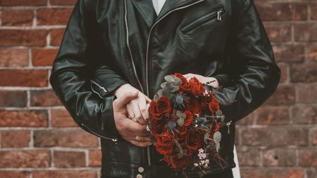 A close-up of a couple standing against a brick wall; the man wears a black leather jacket, and their hands are clasped together holding a bouquet of deep red roses.