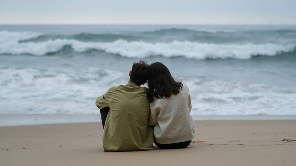 A couple sitting side-by-side on a sandy beach gazing out at the large waves breaking under an overcast sky.