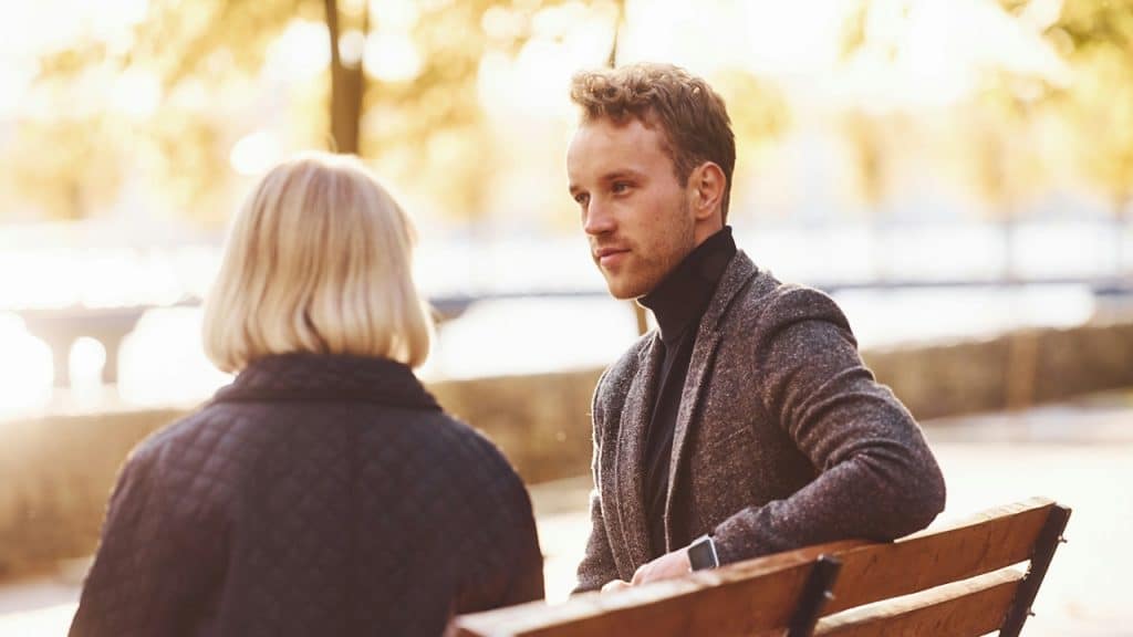 A young man in a gray blazer and black turtleneck sitting on a park bench with a woman.