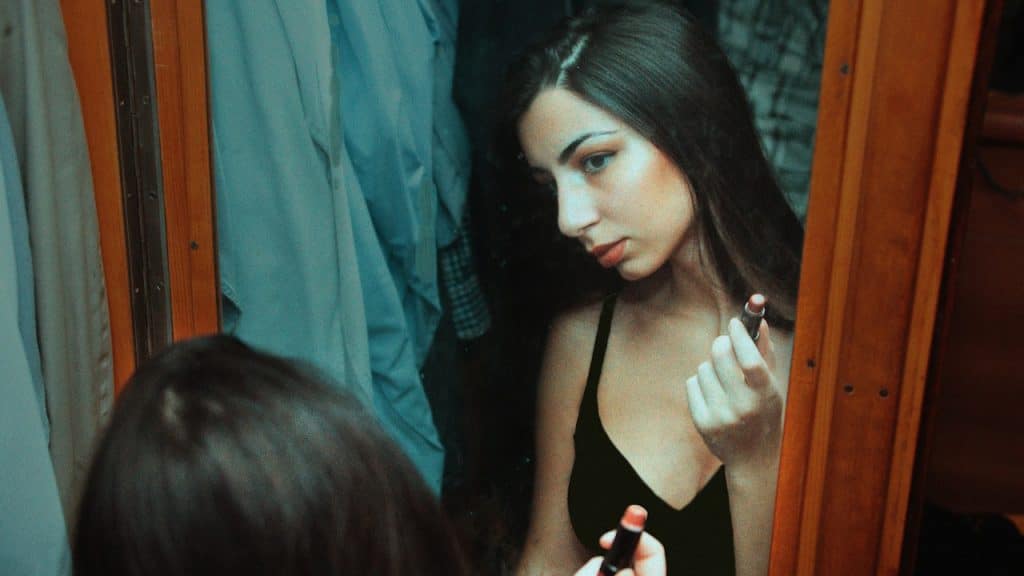 A young woman in a black tank top looking into a mirror while applying red lipstick