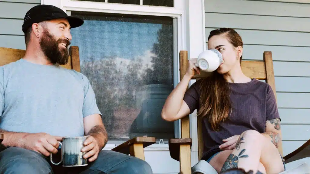 A man and a woman sitting in wooden rocking chairs on a porch, drinking from mugs.