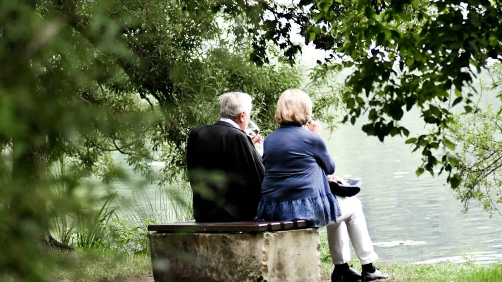 An old couple sitting on a bench by the lake in the woods.
