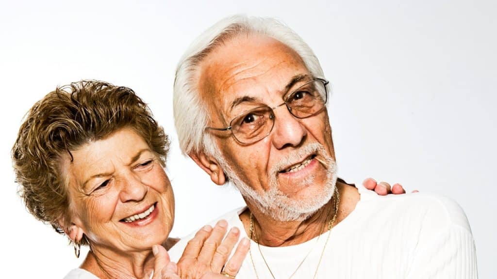An older couple with the woman hugging the bespectacled man from behind.