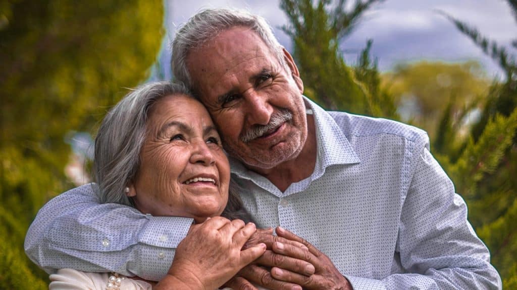 An older man hugging his wife from behind while standing in a field.