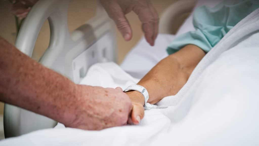 An old man holding his sick wife’s hand in the hospital.