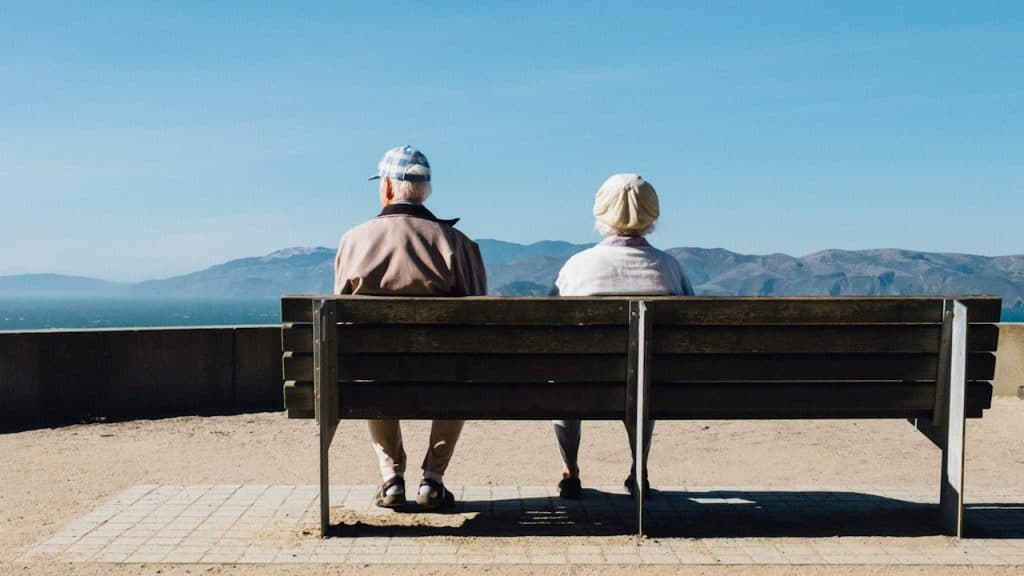 An older couple sitting apart on a bench while gazing at a picturesque view of the mountains.