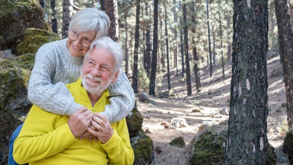An older couple relaxing in the outdoors with the woman hugging the man from behind and him holding her hands