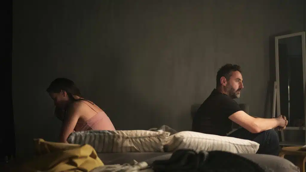 A woman and a man sit facing away from each other on a bed in a dark room.