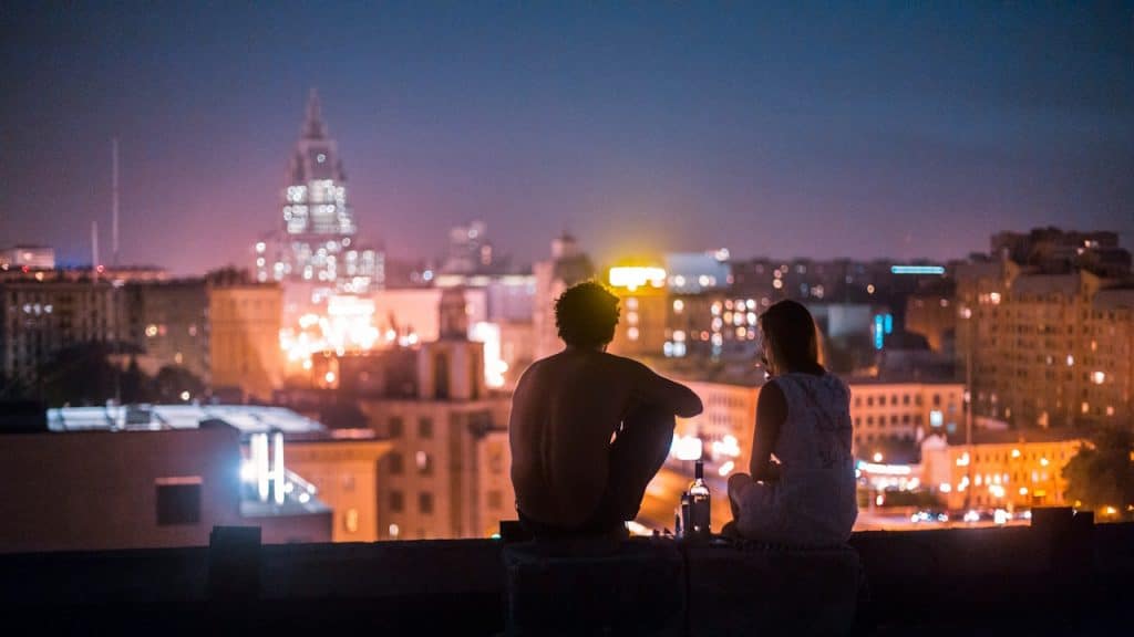 A couple sitting on a rooftop at night looking out over a brightly lit city skyline.