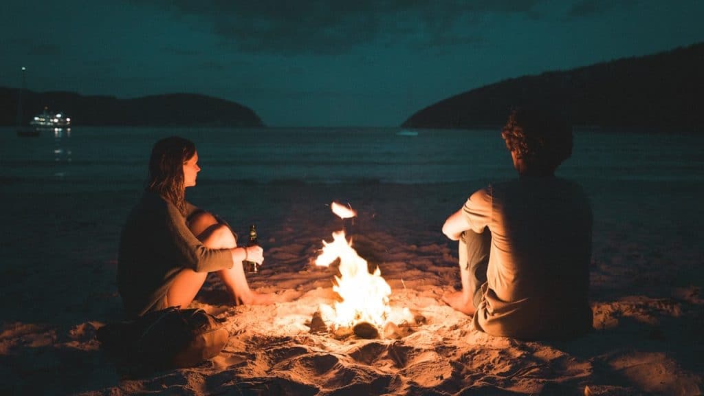 A man and a woman sit facing each other on a sandy beach at night, illuminated by a central campfire.