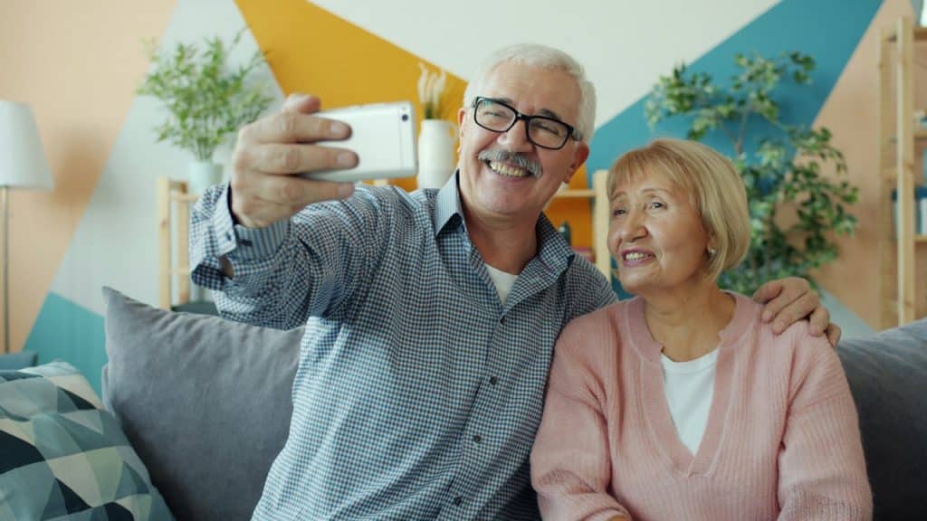 An elderly couple sits on a couch, with the man holding a smartphone up to take a selfie.