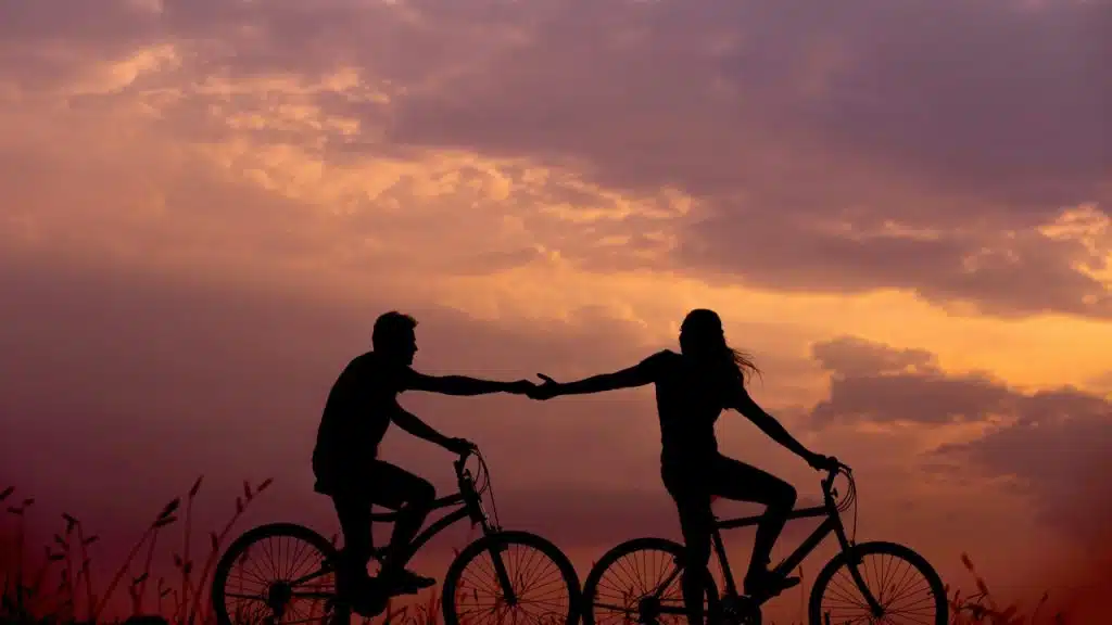 A couple riding bicycles against a fiery sunset sky reaching out to hold hands.