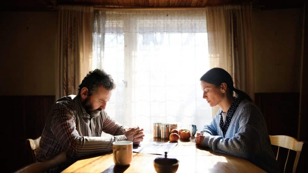 A man and a woman sit at a table in a dimly lit room with their heads bowed and hands clasped.