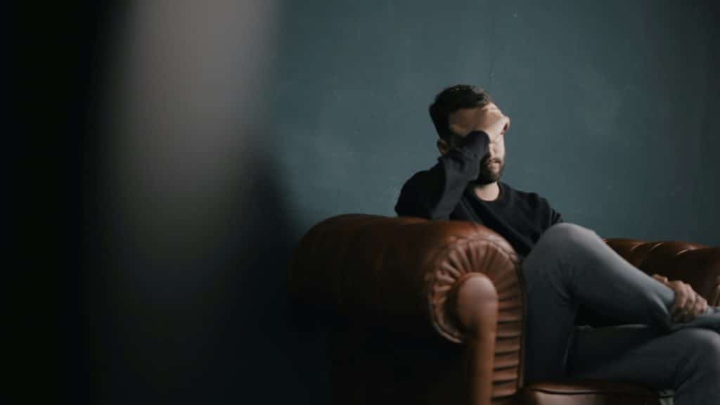 A distressed man in a black shirt and jeans sits on a leather armchair, holding his hand to his forehead in a dimly lit room.