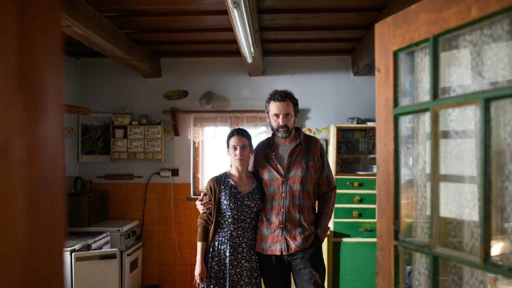 A solemn-looking middle-aged couple stands in a brightly lit and rustic kitchen.