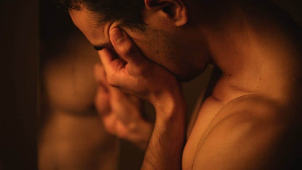 A close-up of a shirtless, muscular man with his hands pressed to his face.