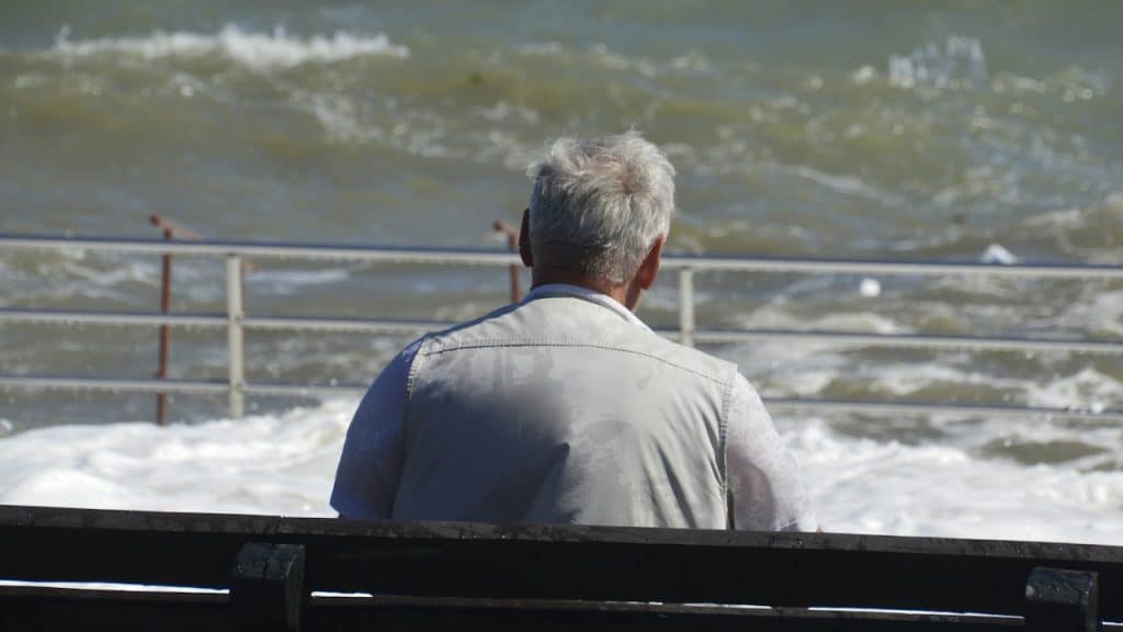 An elderly person with white hair, sits on a dark wooden bench, looking out at the ocean.