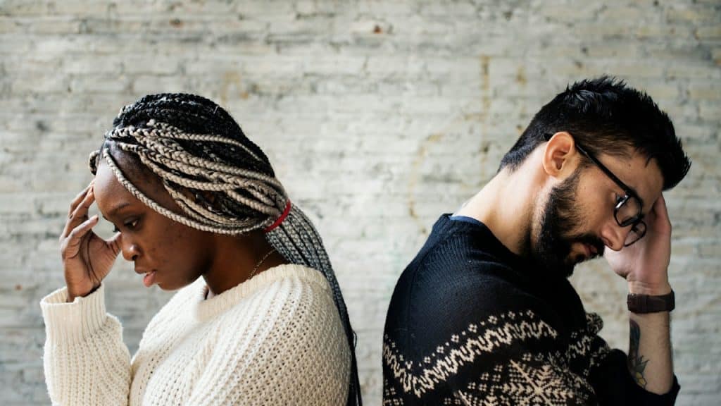 A man and a woman stand back-to-back against a white brick wall, both looking distressed with their hands on their foreheads.