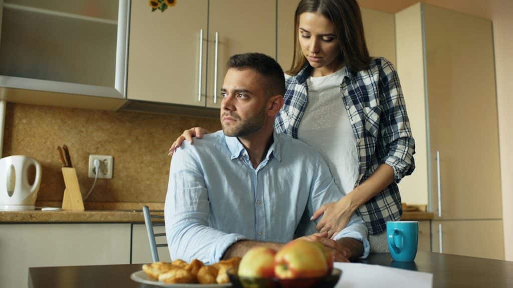 A concerned woman in a plaid shirt stands behind a sullen man in a light blue shirt, resting her hand on his shoulder.