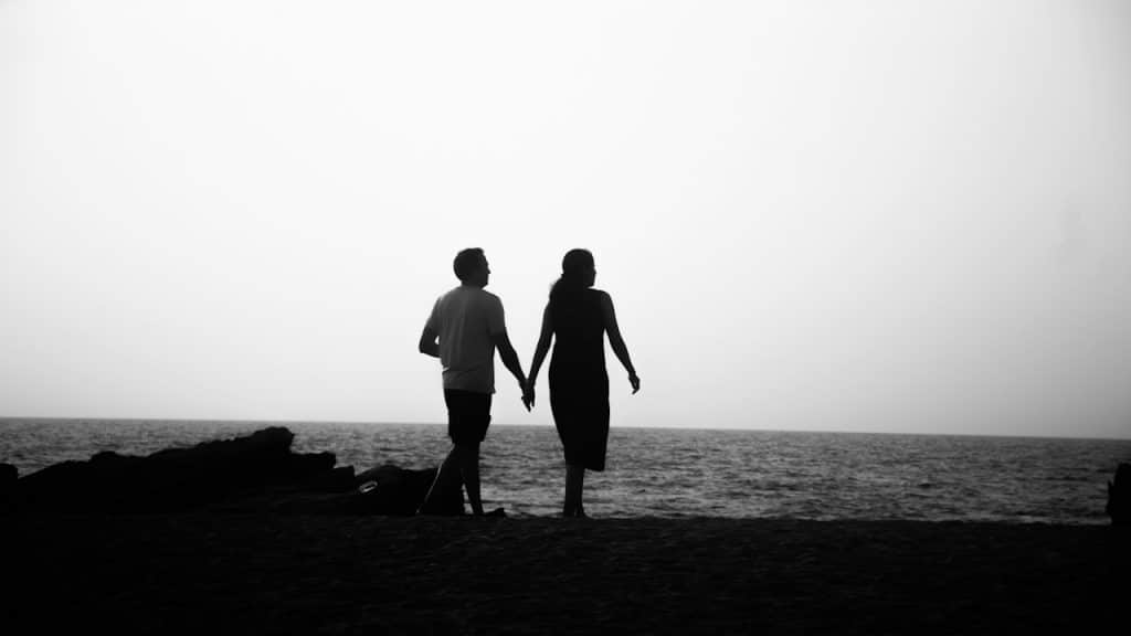A black and white silhouette of a couple holding hands and walking along a beach toward the ocean under a bright, clear sky.