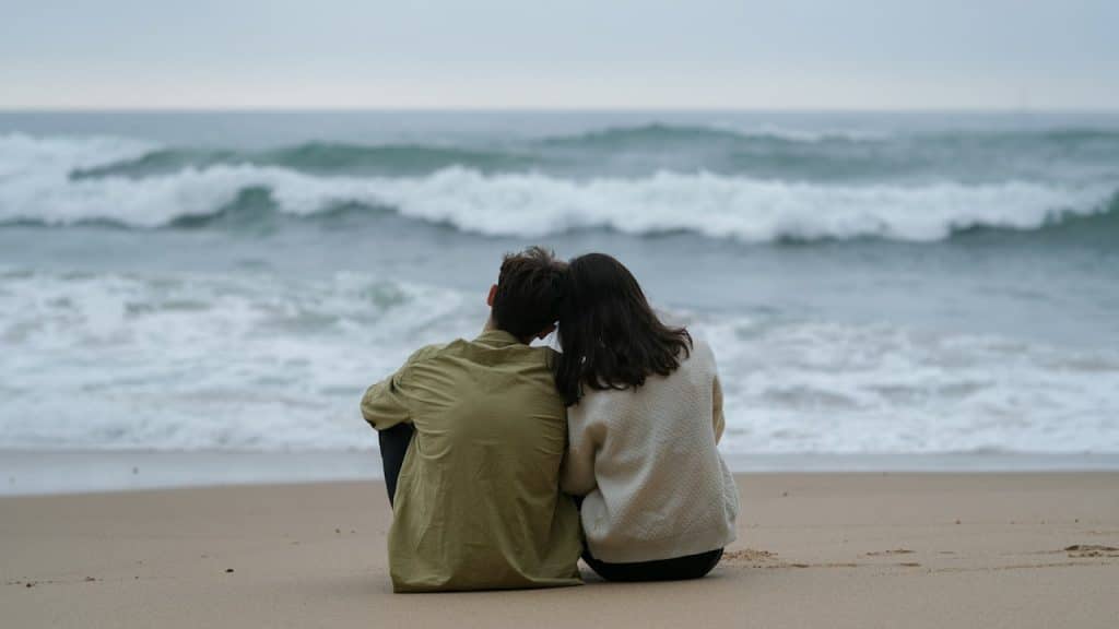 A black and white silhouette of a couple holding hands and walking along a beach toward the ocean under a bright, clear sky.