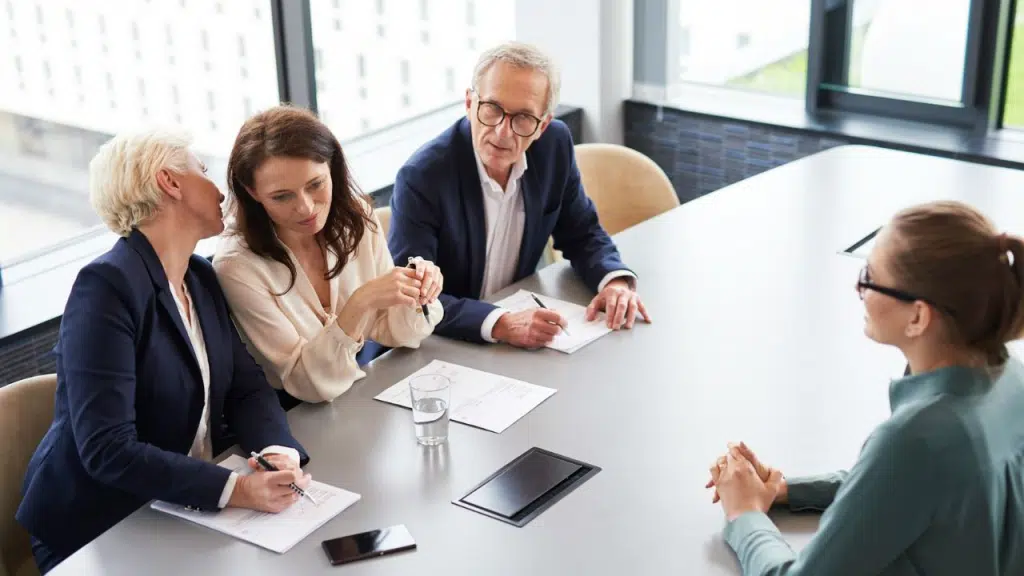 Four professionals, three seated on one side of a conference table, look at a fourth.