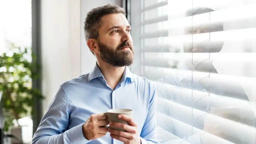A bearded man in a light blue shirt holds a mug and looks thoughtfully out a window with blinds.